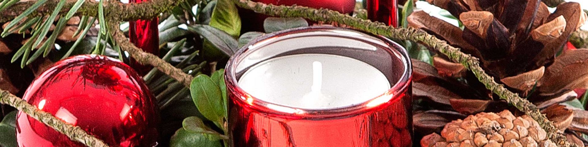 Et dekorativt arrangement med en rød lysestage omgivet af grangrene, kogler og røde julekugler. Stemningen er hyggelig og festlig, der fremhæver julens dekorationer.